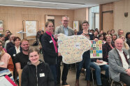3 Personen stehen zwischen von Personen besetzten Stuhlreihen in einem Saal.